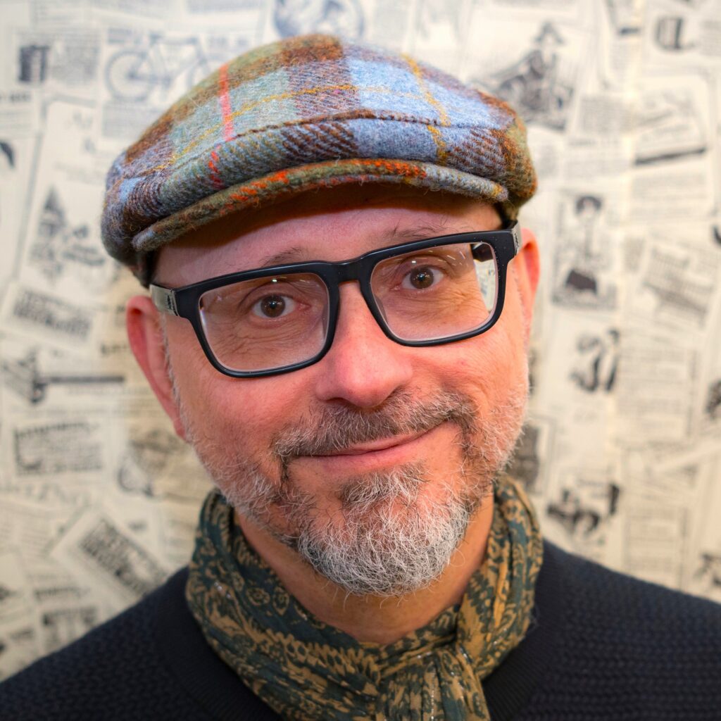 Close-up of a man wearing a blue plaid tweed flat cap and scarf, with black glasses and a newspaper-patterned background.