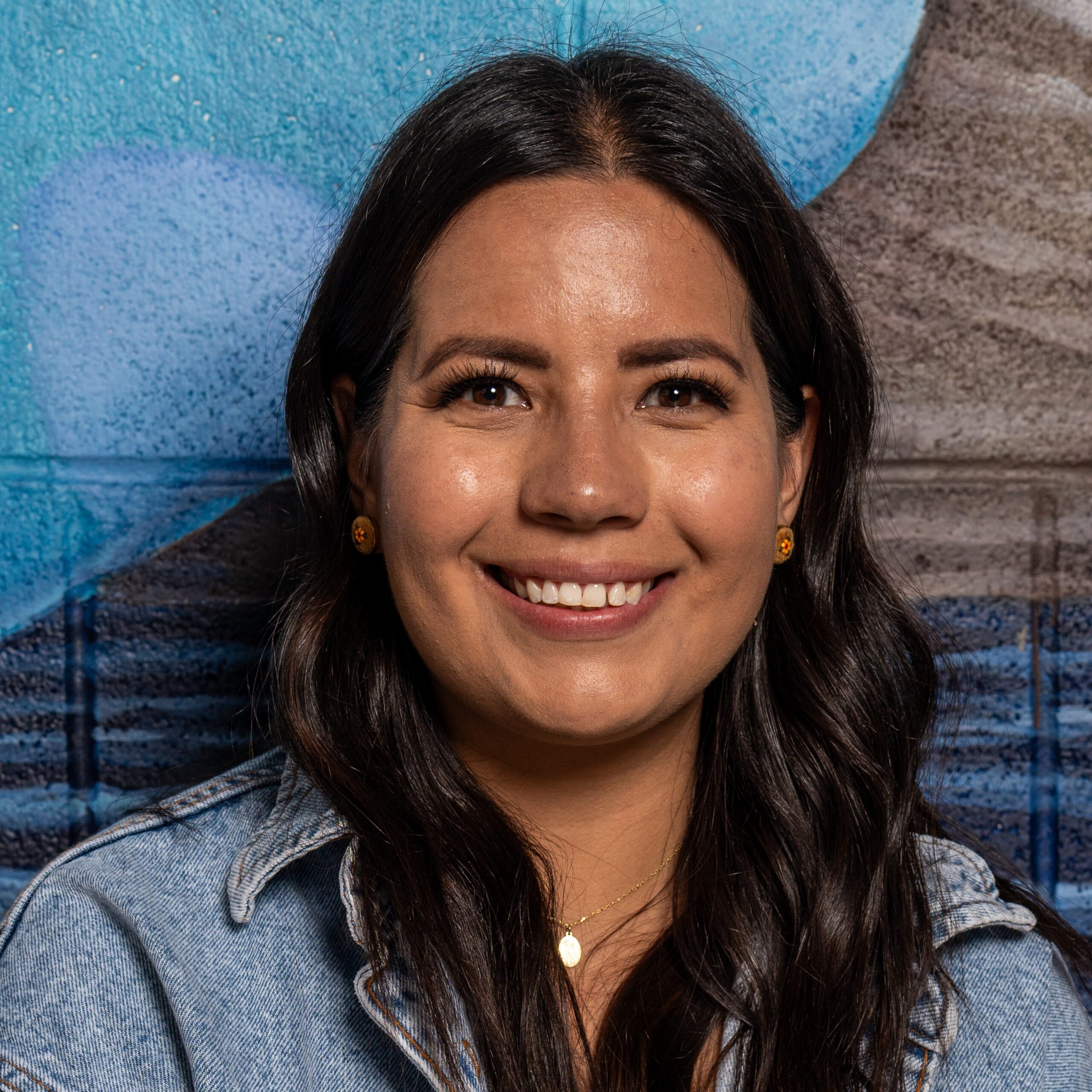 Close-up of a smiling woman with long dark hair wearing a denim jacket against a blue graffiti wall.